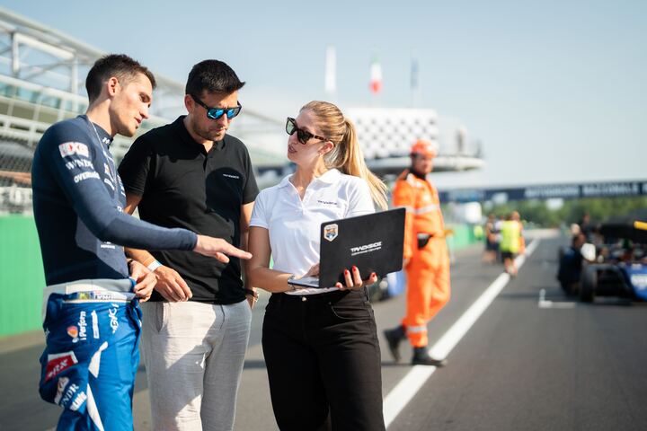 Trackside Command Center crew and a driver reviewing a laptop on the grid at Monza, race cars lined up down the pit straight.